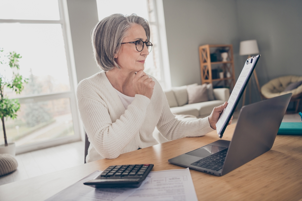 A sophisticated woman in her late 50s wearing a white blazer, thoughtfully reviewing financial documents on a digital tablet in a bright, modern interior.
